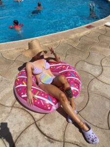 Maryrose, in her bikini and sunhat lounges on a pink donut-shaped pool float near the edge of a swimming pool, confidently enjoying the moment despite having a stoma.