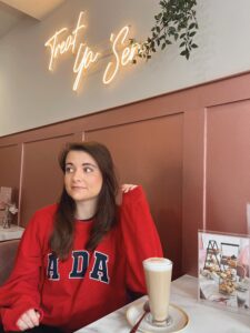 Amy, wearing a bright red sweater, sits at a table sipping her coffee, looking content.