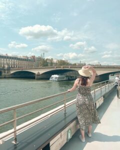 Amy, wearing a dress and hat, leans on a bridge, enjoying the view of the river.