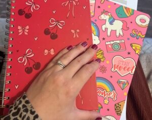 Amy's hand, with painted nails and a ring, appears over her colourful notebooks where she takes notes on her stoma care routine.