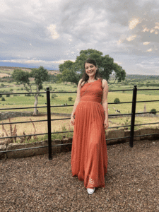 Amy, in a lovely countryside background, smiles wearing a burnt orange dress - proving that you can have confidence with a stoma!