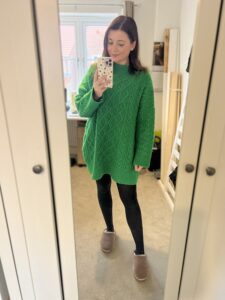 Amy poses in front of her mirror, phone in hand and a smile on her face while she wears a green jumper dress.