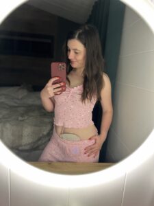 Amy snaps a picture on her phone in her pyjamas, in front of a lit-up mirror, with her stoma bag peaking through her shorts.