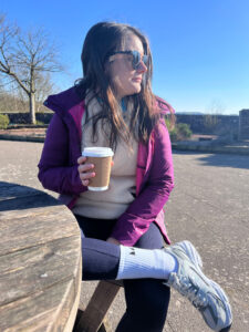 Amy, wearing gym wear, a winter coat and sunglasses, has her picture taken at a picnic bench outside. The sun is shining and she is holding a coffee cup.
