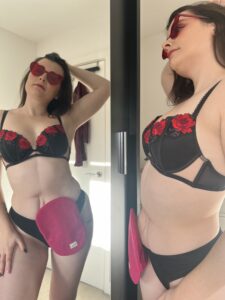 Amy, with heart-shaped sunglasses on, poses in her underwear in front of the mirror. She is also wearing a bright pink stoma bag cover, showing off her confidence.