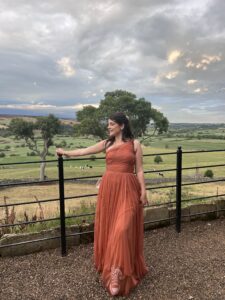 Amy, in front of a natural background, smiles over her shoulder while wearing a burnt orange dress.