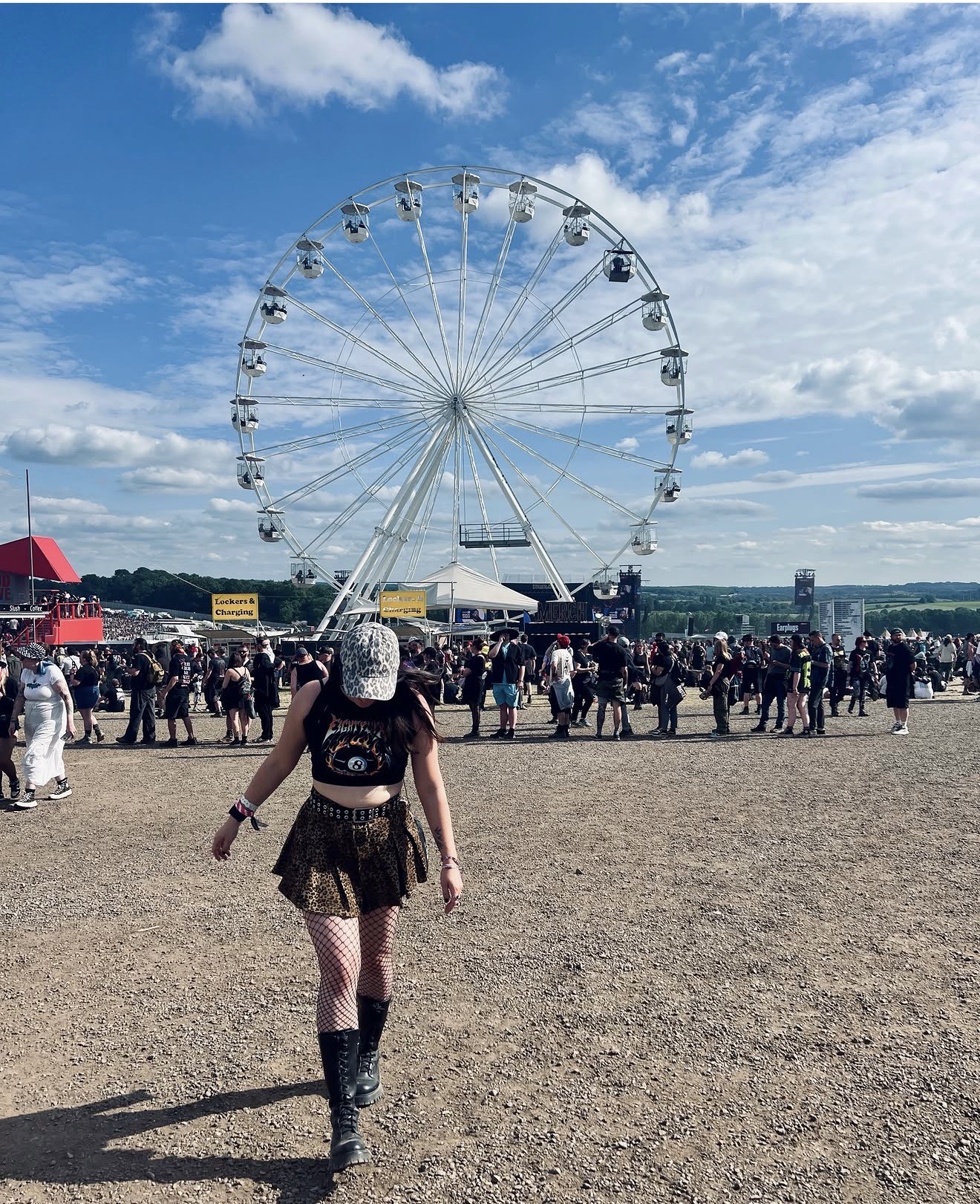 A woman in a hat, black t-shirt and skirt walks in front of a ferris wheel at a busy Download festival