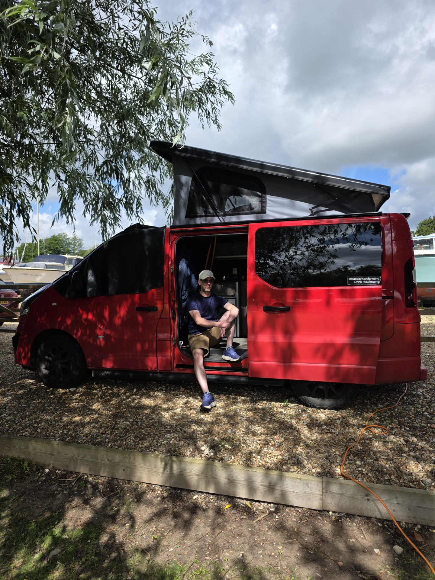 A man sits on the step of his red van, with the pop top extended