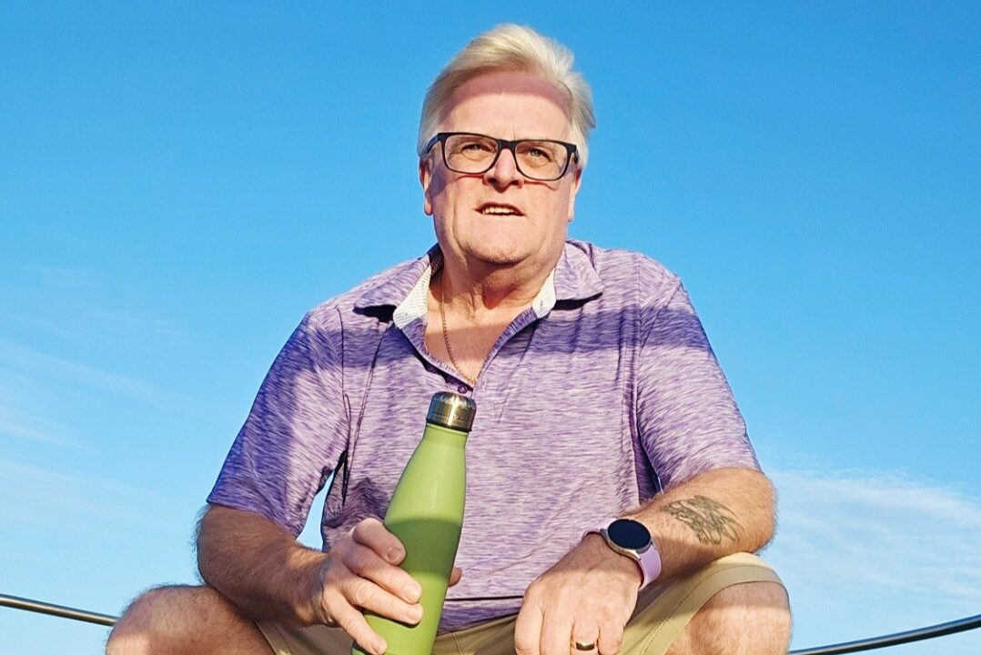 A man squats in front of a clear blue sky with a green bottle in his hand.