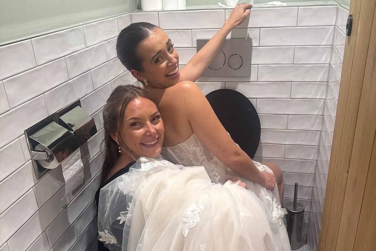 A woman in wedding dress laughs at the camera as she is held her friend, facing the wrong way on the toilet.